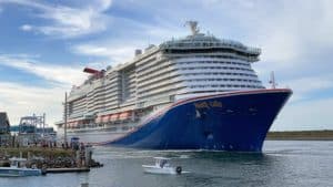 A large cruise ship named "Mardi Gras" from the fleet of Carnival Cruise Line Ships is docked near a harbor. Several smaller boats are in the water nearby, with people onshore observing the scene. The sky is partly cloudy.