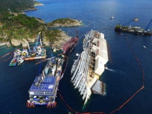 Aerial view of a shipwreck with multiple recovery vessels and cranes surrounding the partially capsized Costa Concordia near a rugged coastline. Brightly colored booms are deployed around the area, and large rocky formations are visible in the background, marking ten years since the disaster.