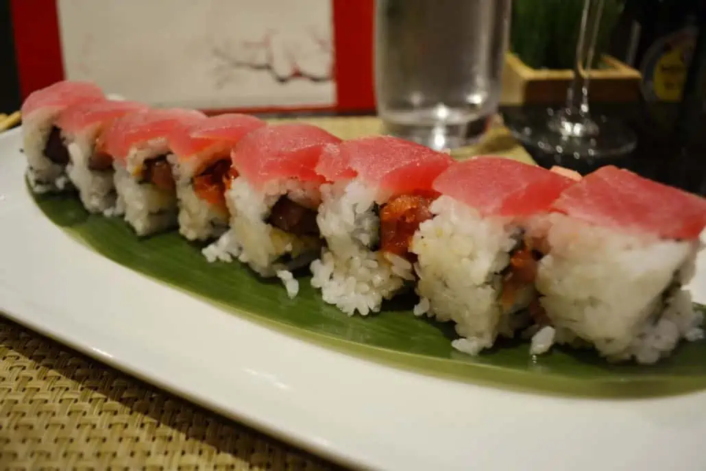 A sushi roll topped with slices of pink tuna is arranged on a rectangular plate lined with a green leaf. A glass of water, a red book, and a small plant are visible in the background, evoking the serenity of an orchid garden. The plate rests on a woven placemat.