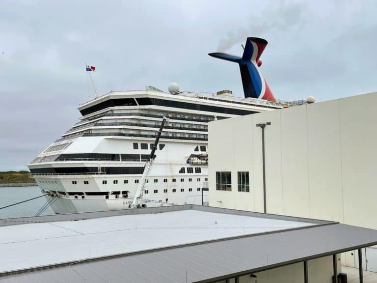 A large cruise ship with a distinctive blue, red, and white funnel is docked at Port Canaveral next to a beige building. The scene is overcast, with grey skies visible in the background, creating an atmospheric backdrop for those setting sail on their adventure.