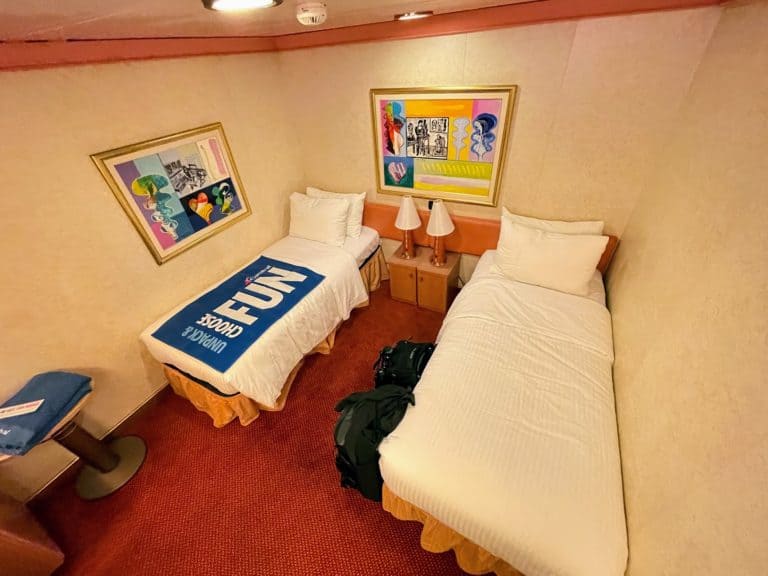 A cruise ship cabin aboard the Carnival Liberty features two single beds with white linens, a nightstand with two lamps between them, and framed abstract artwork above each bed. A luggage bag is on the floor near one bed, and a towel with a logo is draped over the other as you prepare for setting sail from Port Canaveral.