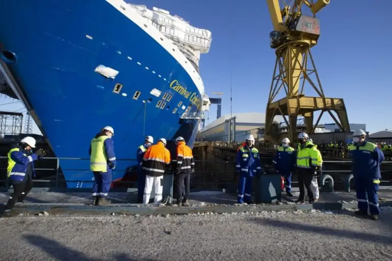 A group of workers in safety gear stand along a dock beside a large blue ship with the words "Carnival Cruise" on its side. A yellow crane is visible in the background, with additional workers and industrial equipment in the vicinity.