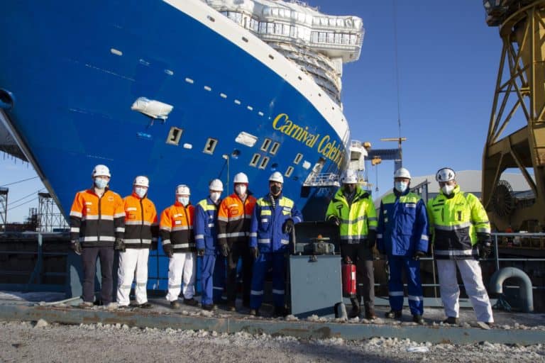A group of nine people in safety gear and helmets stand in front of a large blue ship named 'Carnival Celebration'. The ship is docked and partially visible. The people are dressed in various colors of safety jackets including orange, blue, and green.
