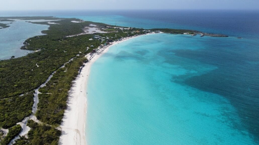 Aerial view of a vast beach with turquoise waters and white sand, bordered by lush green vegetation and small buildings in the distance—one of the must-see sights when exploring Things to Do in Half Moon Cay.