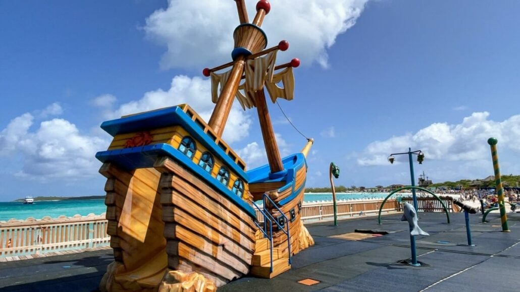 A large wooden pirate ship play structure with a blue sky and ocean in the background, situated beside a playground with various equipment and a crowd of people in the distance. It's one of the delightful things to do in Half Moon Cay.