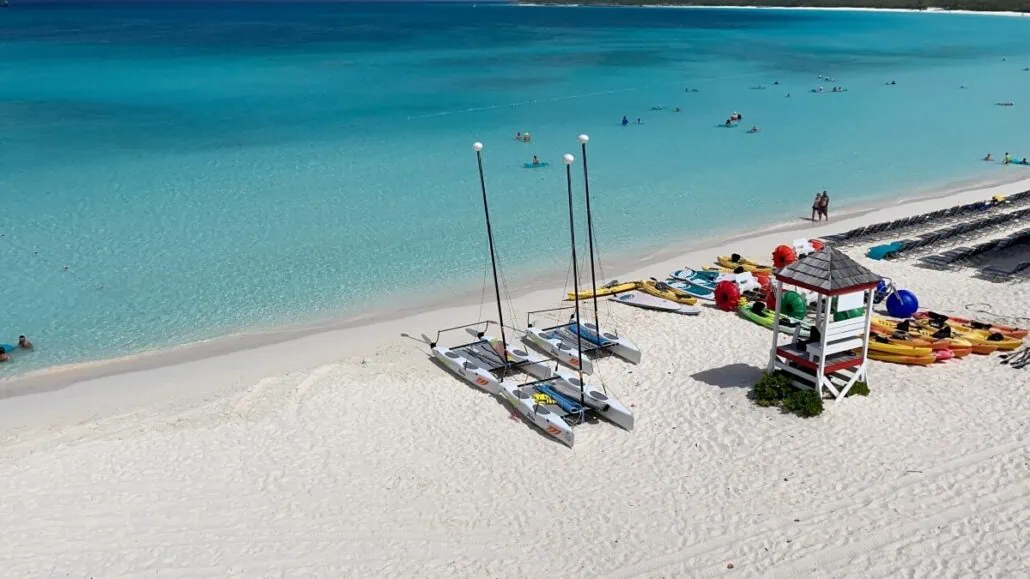 A sandy beach with blue waters, a lifeguard tower, catamarans, kayaks, and beach chairs. People are swimming and relaxing in the background—one of the best things to do in Half Moon Cay.