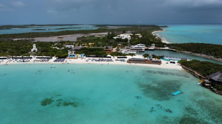 Aerial view of a resort on a coastal area with turquoise waters and a white sandy beach, surrounded by greenery and various buildings. People are visible enjoying the beach, showcasing some of the best things to do in Half Moon Cay.