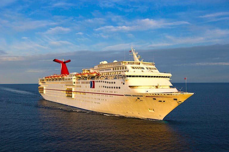 A large white Carnival cruise ship with a red and blue logo is sailing on a calm sea under a clear sky. The ship has multiple decks with rows of windows and lifeboats visible. The sun is casting a golden light on one side of the ship, creating a striking contrast against the serene backdrop.