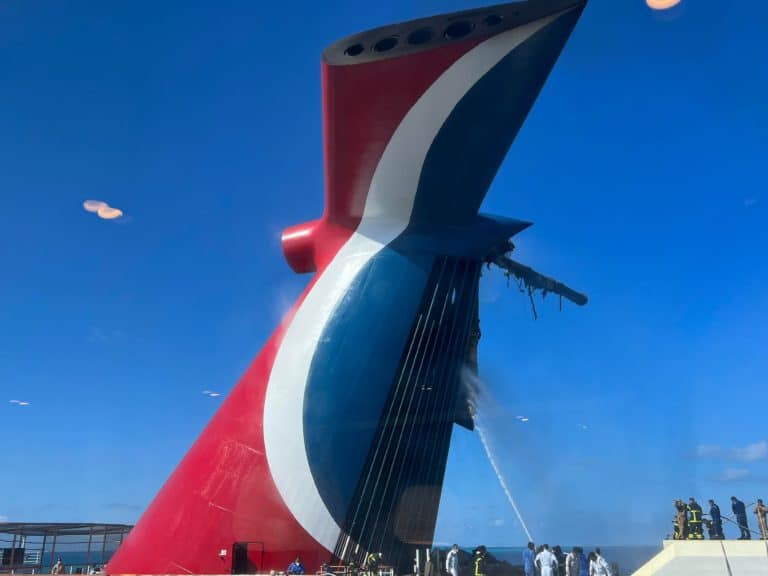 A group of people in safety gear are seen near the distinctive red, white, and blue funnel of Carnival Freedom. Some are using hoses to spray water at the structure, possibly for maintenance or cleaning. The sky is clear and blue, with a few reflections on a glass panel.