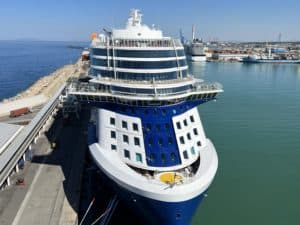 A large blue and white cruise ship, part of the renowned European Cruises, is docked at a pier with calm waters surrounding it. The ship boasts multiple decks with windows and a helipad on the front. In the background, another ship and industrial structures are visible under a clear blue sky.