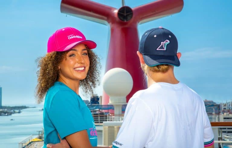 Two people stand on a cruise ship's deck during the Carnival Celebration with a large red funnel in the background. The person on the left smiles at the camera, wearing a pink cap and teal shirt. The person on the right, sporting exclusive merchandise with a blue cap and white shirt, has their back to the camera.