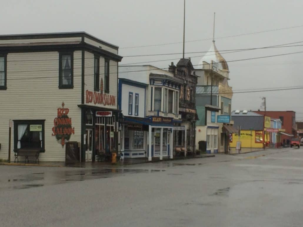 A street scene in the small Alaskan town of Skagway features old-fashioned wooden buildings, including a saloon and various shops. The wet road hints at recent rain, adding to its charm. It's a place brimming with history and beauty, ideal for exploring countless things to do.