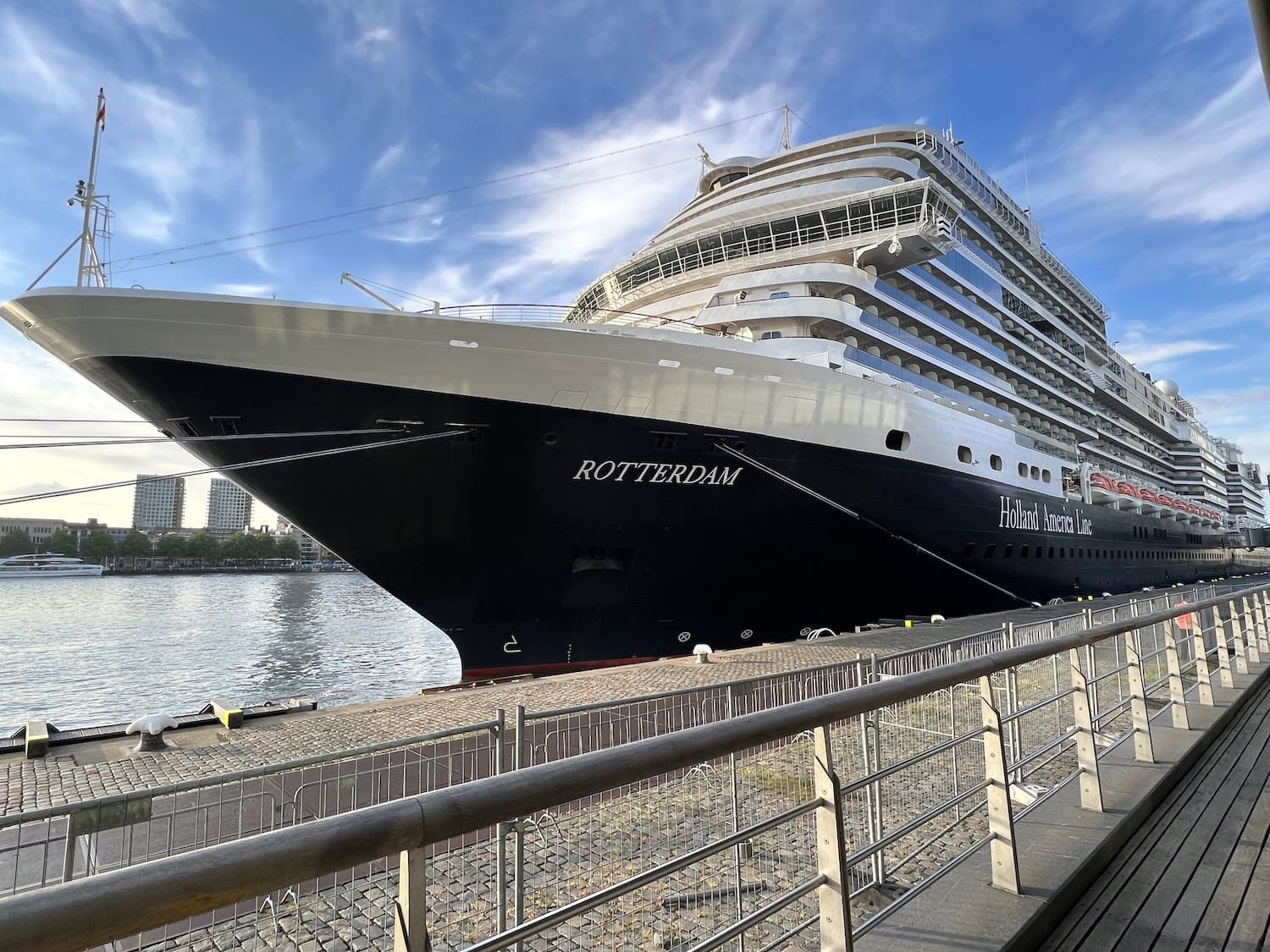 The ms Rotterdam, a grand cruise ship with sleek black and white upper decks, is docked beside a paved walkway. In the background, buildings rise against a partly cloudy sky, complementing the majestic vessel's serene presence.