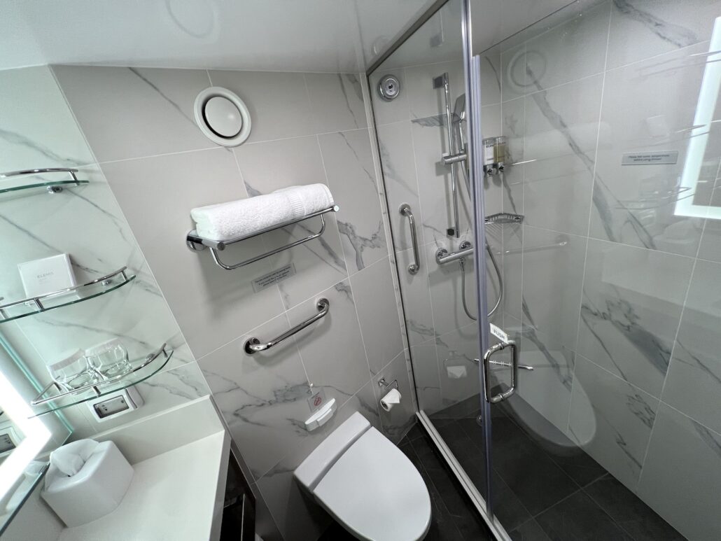 A modern bathroom aboard the ms Rotterdam features a glass-enclosed shower, white toilet, and marble-patterned tiles. A towel is neatly placed on a metal shelf above the toilet. Glass shelves hold toiletries and glasses, with a lit mirror above the sink elevating the elegance.