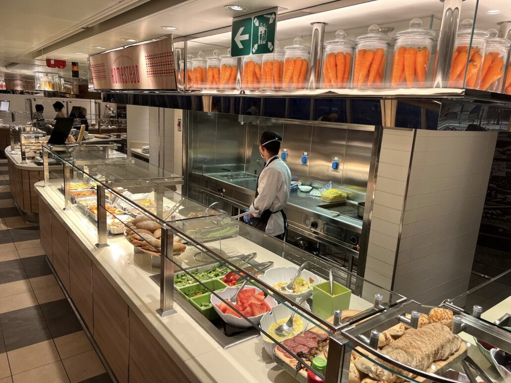 A cafeteria-style buffet on the Ms Rotterdam features an array of foods, including sliced meats, bread, and vegetables. A person in a chef's uniform works diligently behind the counter. Containers of carrots are artfully displayed above the tiled floor.