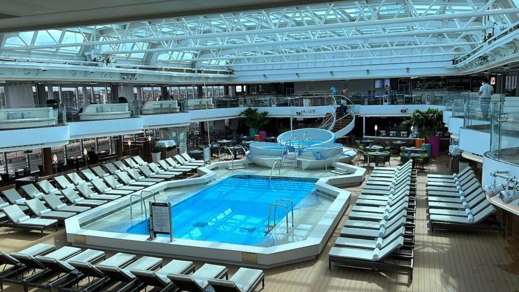 The indoor pool area on the MS Rotterdam boasts a rectangular swimming pool surrounded by numerous lounge chairs. Large skylights illuminate the space, while plants and seating areas grace the upper level, creating an inviting oasis at sea.