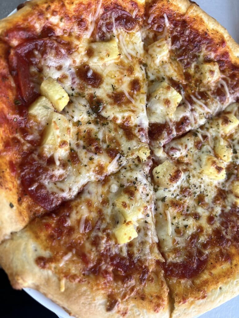 A close-up of a pizza aboard the ms Rotterdam, topped with cheese, pineapple chunks, and pepperoni. The crust is golden-brown, and melted cheese is sprinkled with herbs. Sliced into several pieces, it beckons passengers with its appetizing aroma.