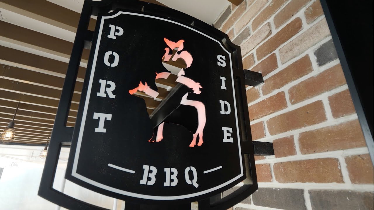 A black and white sign for "Port Side BBQ" evokes cruise line BBQ vibes, featuring a stylized silhouette of a person riding an animal, possibly a pig, with orange flames. The sign is mounted on a wall with exposed bricks.