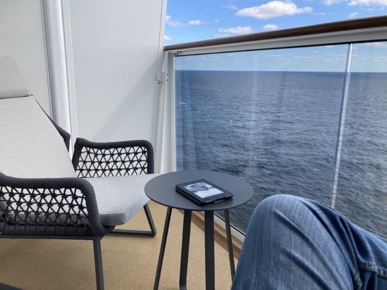 A small balcony on the Norwegian Prima features a chair with a light cushion, a side table with an e-reader, and a man's leg in blue jeans in the foreground. The balcony overlooks a calm sea with a blue sky and scattered clouds.