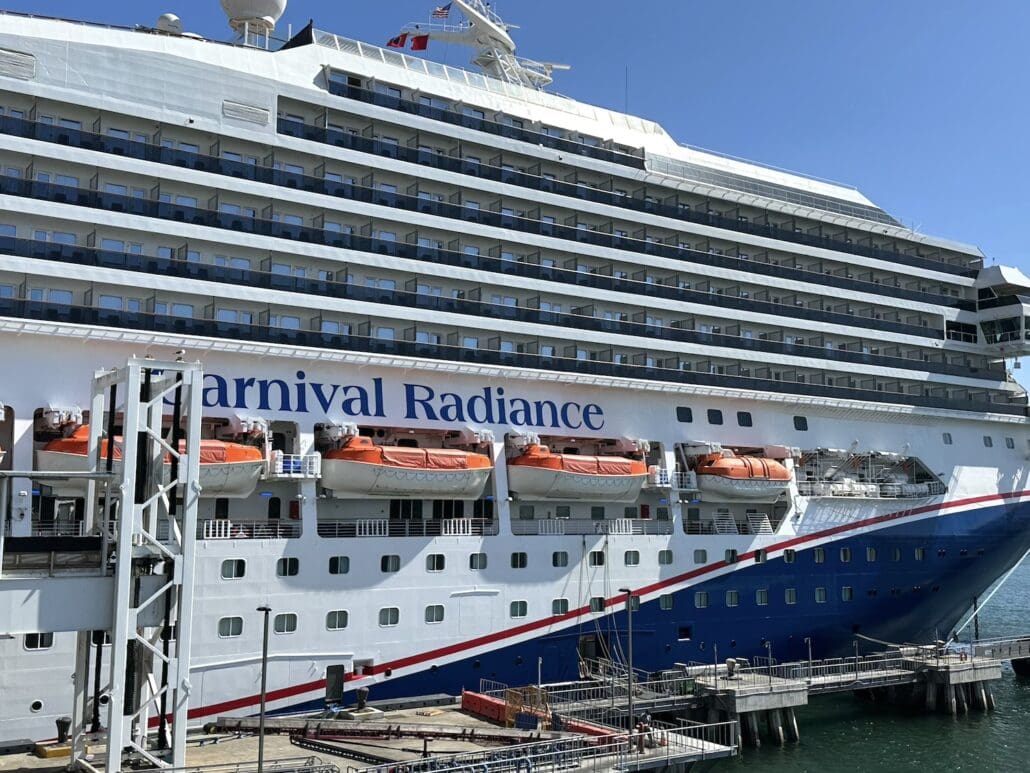 The majestic Carnival Radiance is docked at the pier under a clear blue sky. Multiple decks with rows of windows and vibrant orange lifeboats grace the ship's side, while its hull gleams in blue. For a memorable voyage, explore some insider Carnival Radiance tips before setting sail.