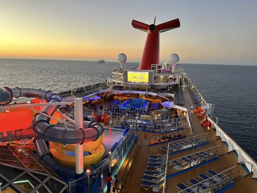 Aboard the Carnival Radiance, the cruise ship deck at sunset offers a spectacular scene featuring a water slide and numerous lounge chairs. A large screen entertains those seated around tables. The serene waters make this moment magical, as a distant ship dots the horizon—truly an unforgettable journey.