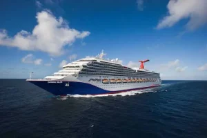 A large Carnival Cruise Ship named "Carnival Valor" is sailing on the ocean under a clear blue sky with a few scattered clouds. The ship features multiple decks, lifeboats, and a distinctive red and blue funnel with the Carnival logo, ready for any rescues in collaboration with the Coast Guard.