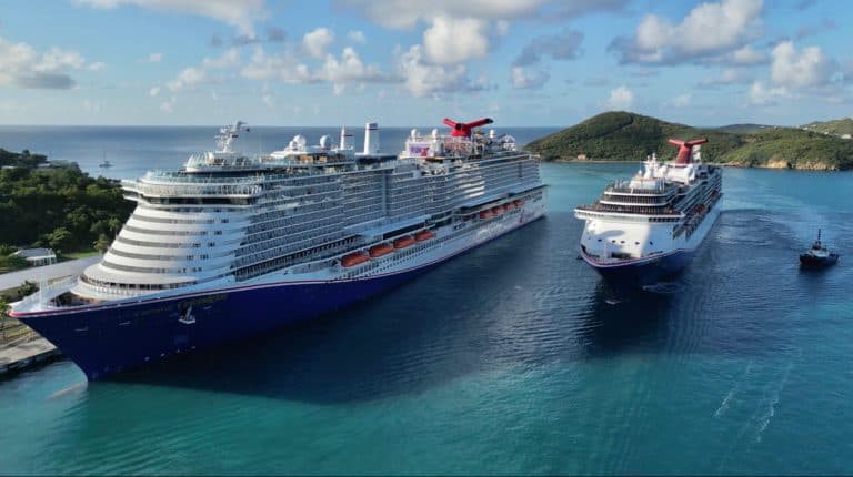 Two large cruise ships are docked side by side in a turquoise bay near a lush, green island. A smaller boat is seen near the ship on the right. The sky is partly cloudy with a mix of white and gray clouds.