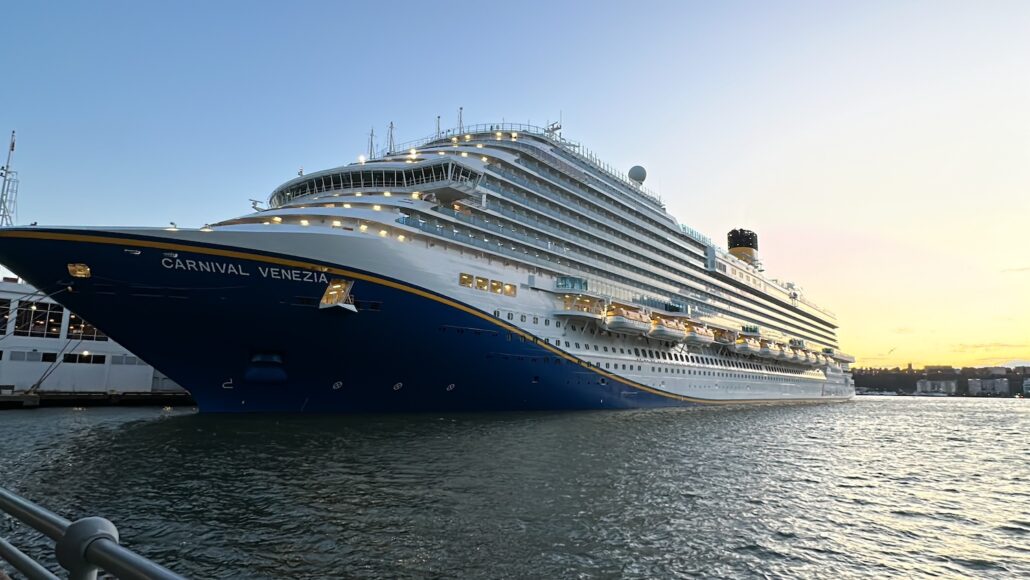 As the sun sets, the majestic Carnival Venezia docks at port, its illuminated decks and windows casting a brilliant glow. The evening light dances on the water, enhancing the ship's grandeur.