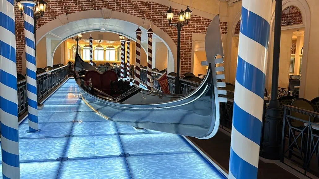 A shiny gondola is displayed indoors on a bridge over a glowing blue floor, evoking the charm of Carnival. The scene, perfect for Venezia dining, is framed by striped poles and surrounded by a warm, yellow-lit interior with brick accents and arched windows in the background.
