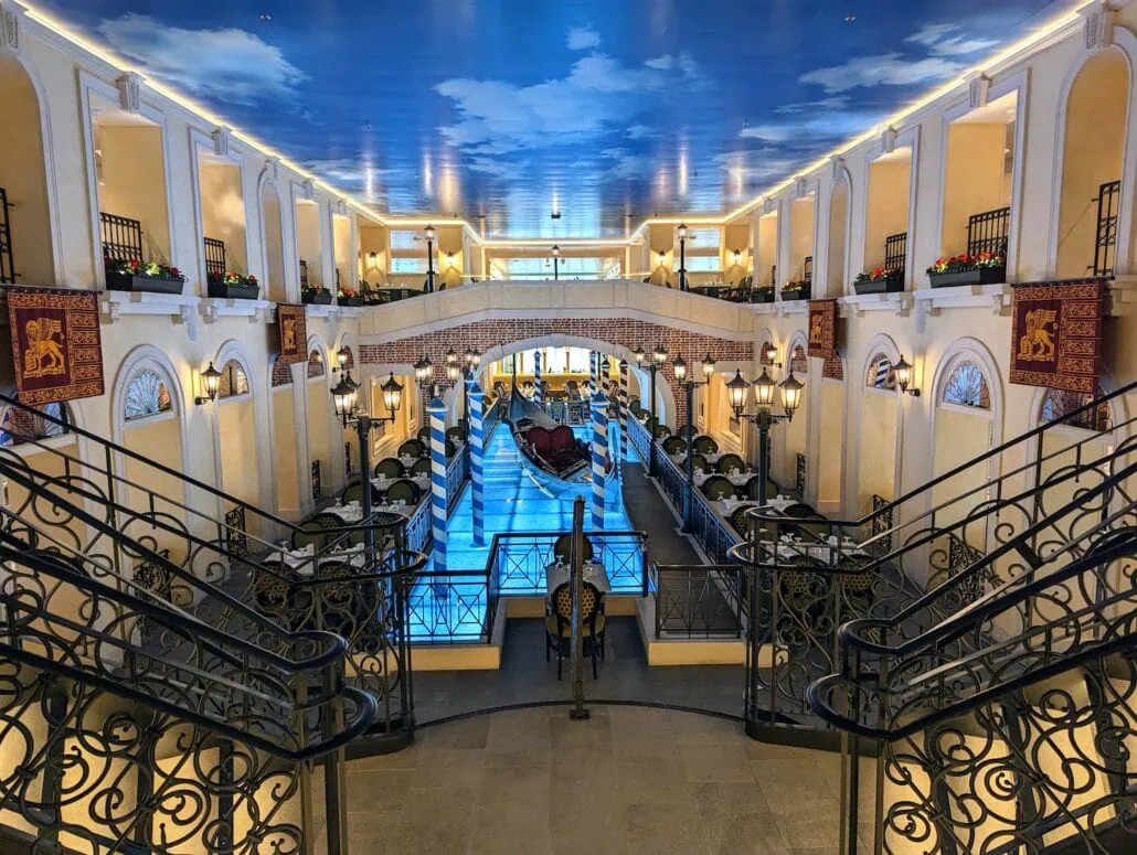 View of an ornate indoor space resembling a Venetian canal scene, featuring railings, a painted sky ceiling, and a gondola in a waterway. Various levels with arches and small balconies add to the luxurious atmosphere, reminiscent of the opulence found in Carnival Venezia dining experiences.