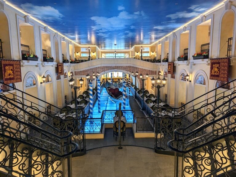 View of an ornate indoor space resembling a Venetian canal scene, featuring railings, a painted sky ceiling, and a gondola in a waterway. Various levels with arches and small balconies add to the luxurious atmosphere, reminiscent of the opulence found in Carnival Venezia dining experiences.