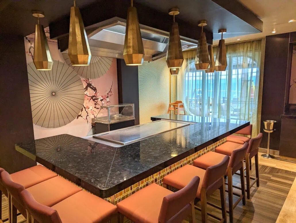 The Carnival Venezia Dining experience offers a teppanyaki grill setup with a sleek black marble countertop surrounded by cushioned chairs. Brass pendant lights hang overhead, while the artistic backdrop showcases umbrellas and cherry blossoms. Large windows flood the space with natural light.