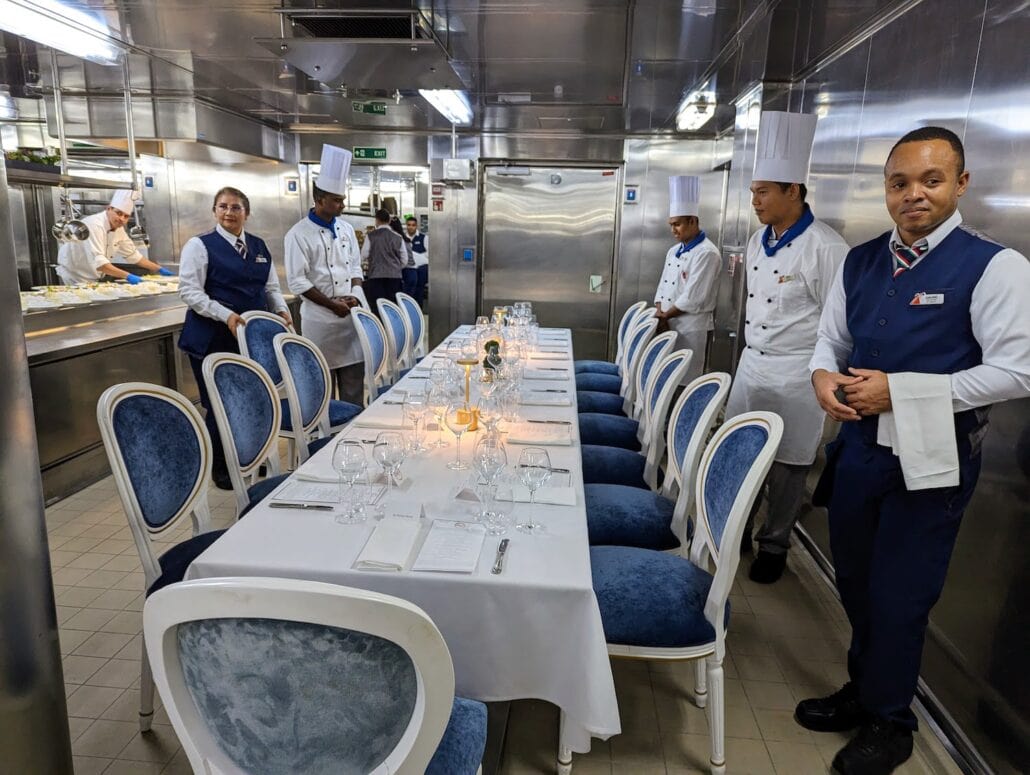 A group of five restaurant staff members in uniforms stands next to a Carnival Venezia dining table set for 10, with a white tablecloth, blue chairs, and neatly arranged glassware in a stainless steel kitchen setting.