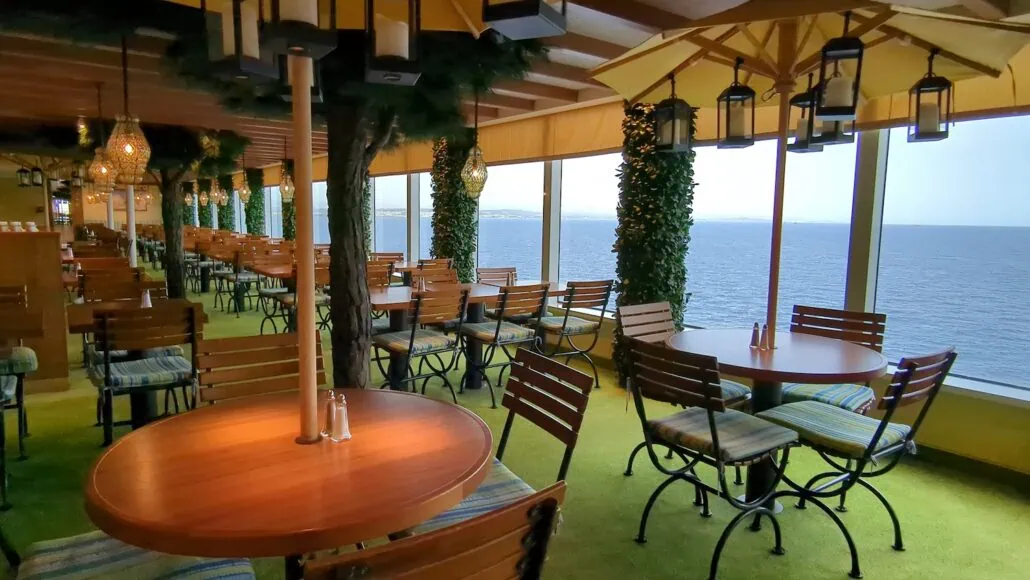 Carnival Venezia Dining offers a unique experience with wooden tables and chairs set on a lush green carpet. Large windows frame stunning ocean views, while elegant ceiling lights illuminate the space adorned with vibrant plants and trees.