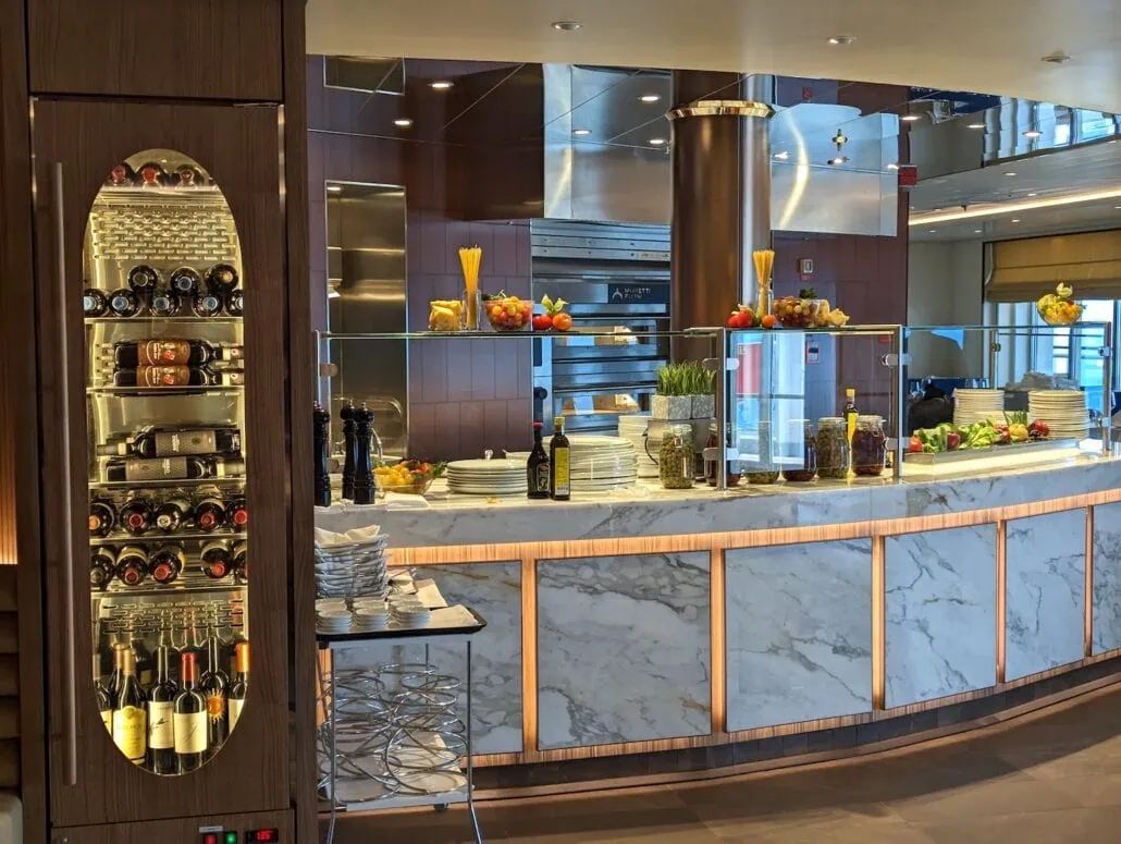 A modern kitchen with a marble counter, showcasing Carnival Venezia dining flair through bottles of olive oil, wine, and stacks of plates. The counter also features glass jars and bowls of fresh produce like peppers and lemons. A wine rack is embedded in the dark wood cabinetry on the left.