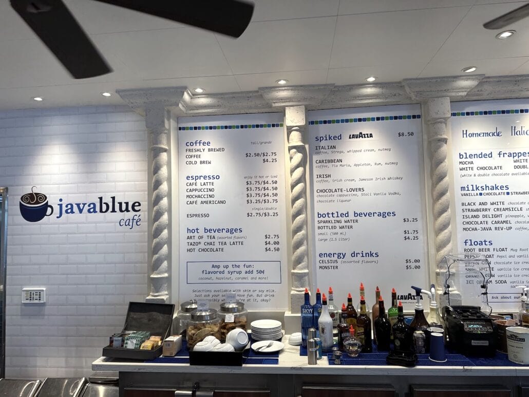 A café counter with various beverages listed on three large menu boards evokes the vibrant atmosphere of Carnival Venezia dining. The options include coffee, spiked drinks, bottled beverages, and energy drinks. The counter displays syrups, jars, and a milkshake machine at Javablue Café.