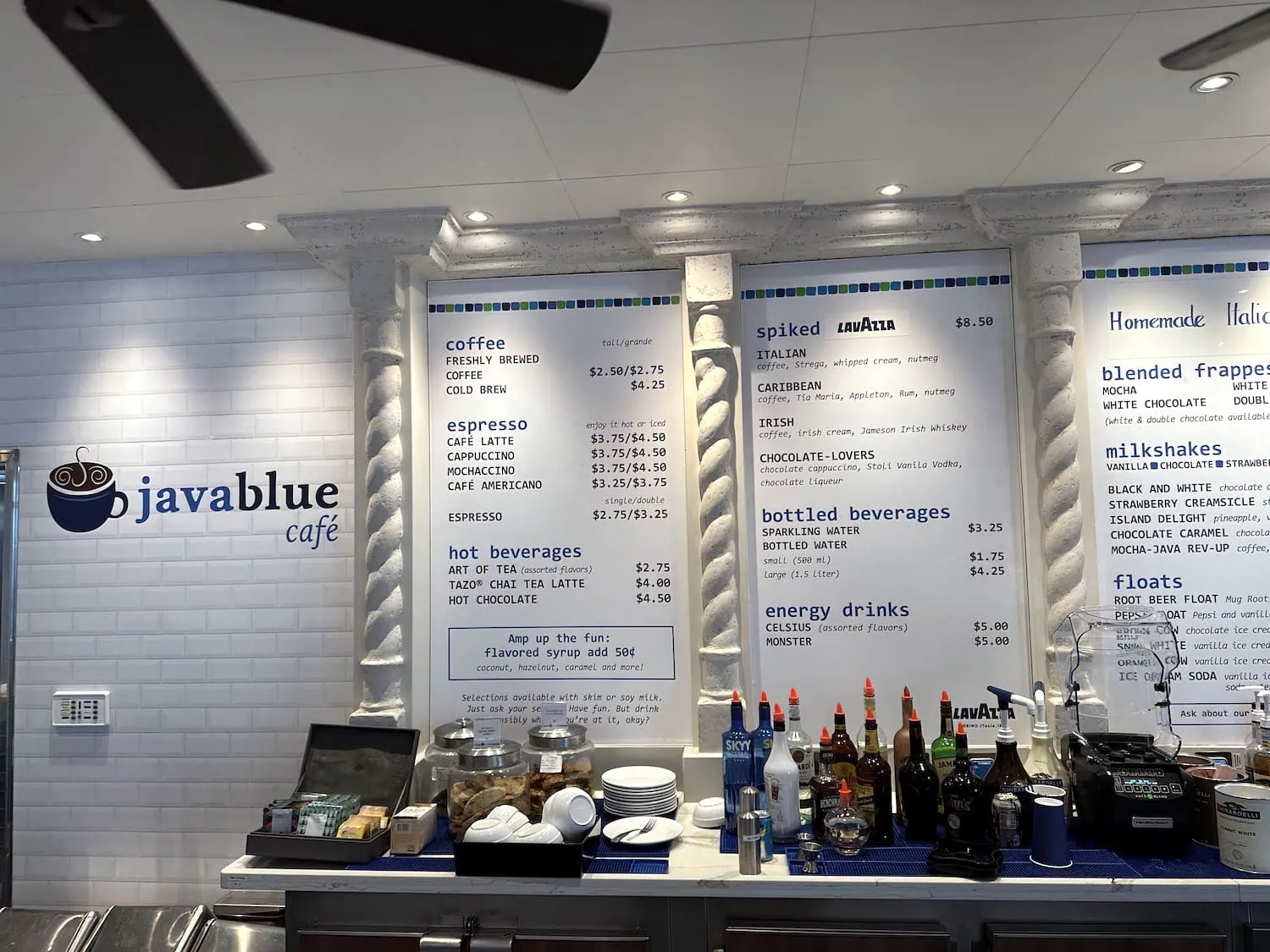 A café counter with various beverages listed on three large menu boards evokes the vibrant atmosphere of Carnival Venezia dining. The options include coffee, spiked drinks, bottled beverages, and energy drinks. The counter displays syrups, jars, and a milkshake machine at Javablue Café.