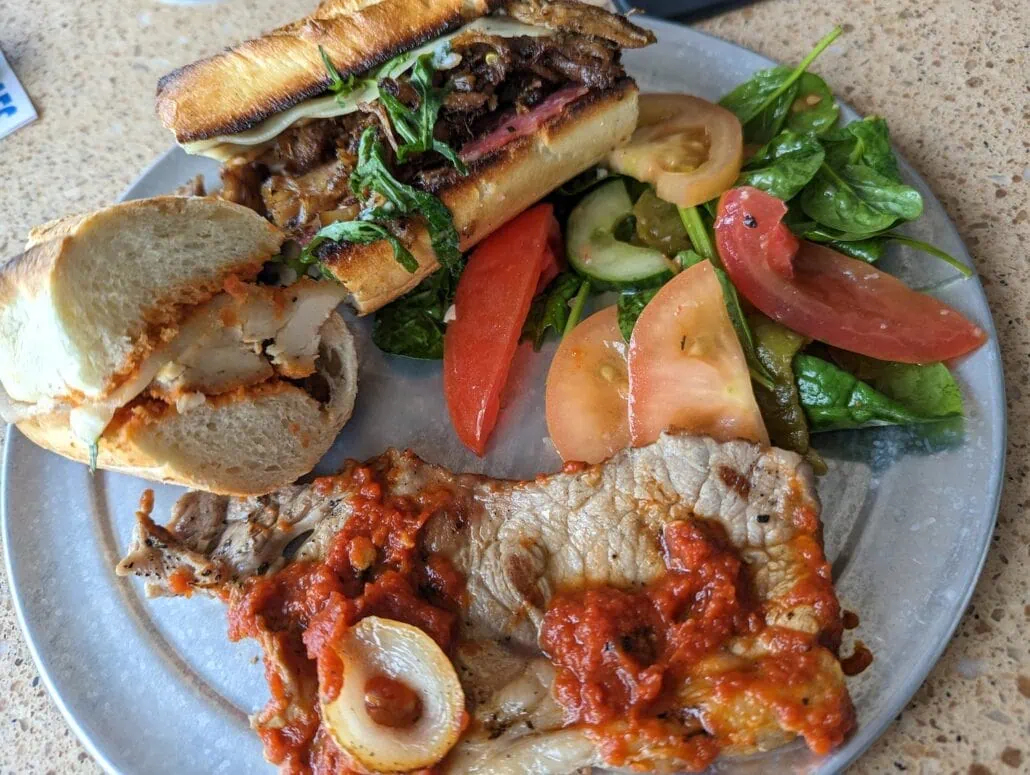A plate showcasing the Carnival Venezia dining experience: a toasted sandwich filled with savory meat and herbs, a fresh salad of tomatoes, cucumbers, and greens, and a piece of grilled meat topped with red sauce and sliced onions.