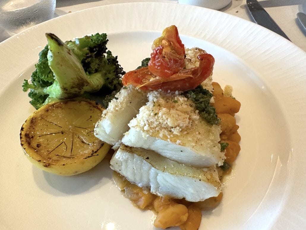 A plate of white fish fillets topped with a breadcrumb crust and roasted cherry tomatoes, reminiscent of Carnival Venezia dining, served with grilled broccoli, a lemon wedge, and yellow beans in a savory sauce.