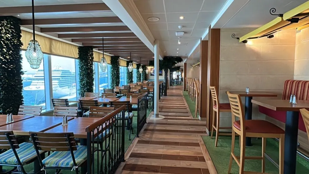 The restaurant interior features wooden tables and striped cushioned chairs, exuding a Carnival Venezia dining vibe. Greenery and lantern-style lights enhance the ambiance, while large windows reveal captivating views of a cruise ship. The floor combines wood with green carpeting for added charm.