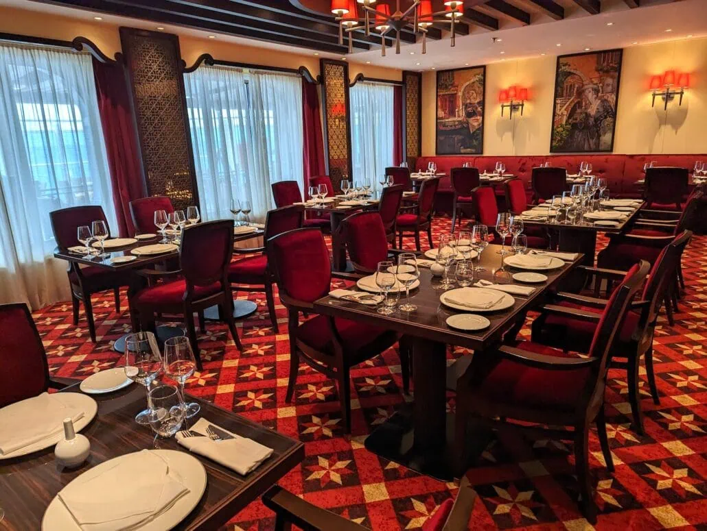 The Carnival Venezia Dining area showcases red upholstered chairs around dark wooden tables adorned with wine glasses and elegant table settings. The room's patterned red carpeting, window curtains, and wall sconces contribute to its formal and sophisticated ambiance.