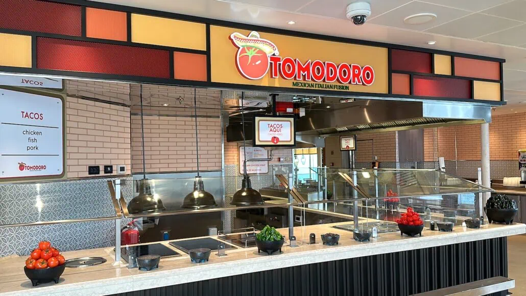 A Mexican dining station named "Tomodoro" aboard Carnival Venezia, featuring a vibrant sign. The counter showcases bowls of red and green peppers, with a menu offering tacos with chicken, fish, or pork options. Kitchen equipment is visible in the background.