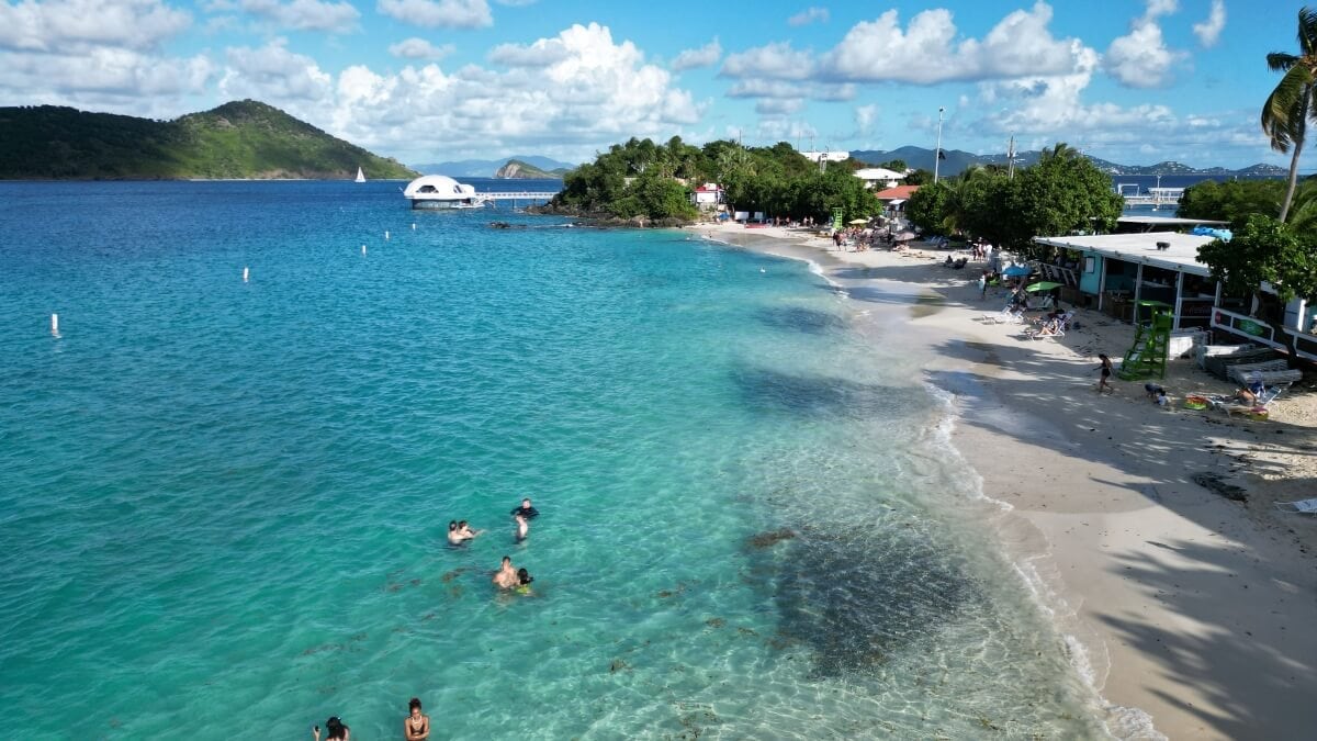 A tropical beach scene with clear blue water, people swimming, a sandy shore, and buildings on the right. Trees line the coast of one of the beautiful beaches in St. Thomas. A small island and more water are in the background under a partly cloudy sky.