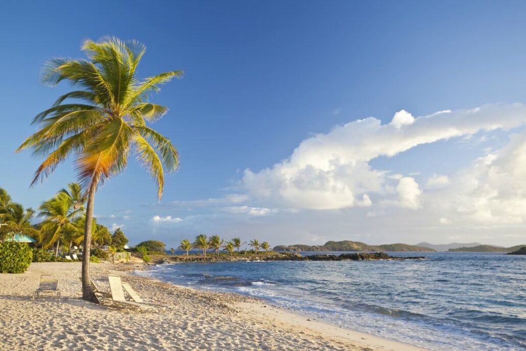 A tropical beach with white sand, palm trees, and clear blue water under a partly cloudy sky. Two lounge chairs are placed under a palm tree. Small islands are visible in the distance—truly one of the idyllic beaches in St. Thomas.
