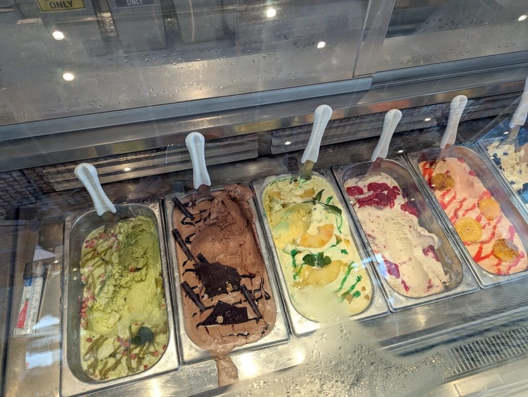 A display case reminiscent of Carnival—with trays of delectable gelato delights, echoing the vibrant colors of Venezia. Each tray, adorned with a white plastic spoon, showcases flavors in green, brown, light yellow, and mixed hues topped with chocolate pieces and fruit.