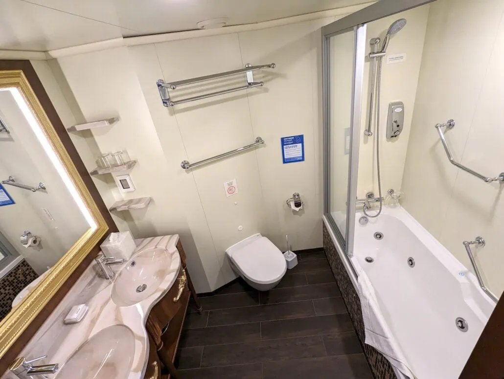 A bathroom reminiscent of a Carnival Venezia cabin, featuring a double sink with a marble countertop, a wall-mounted mirror, and a shower-bathtub combo with sliding glass doors. The floor is tiled in dark tones and several towel racks are mounted on the walls.