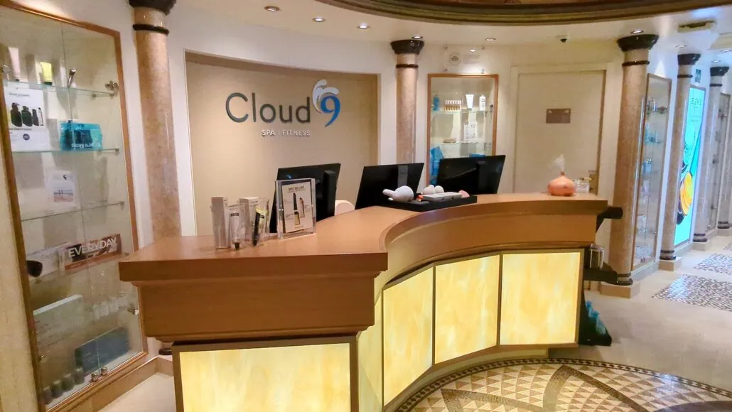 The reception area of Cloud 9 Spa Fitness exudes elegance with its curved wooden desk and backlit paneling, reminiscent of the luxurious Carnival Venezia cabins. Display shelves showcase premium products and decor while two computer monitors sit ready on the desk.