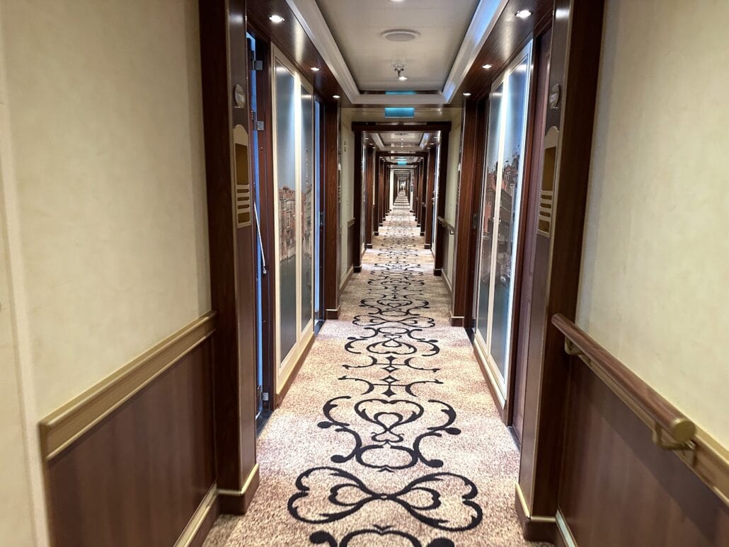 A well-lit hallway extends with a patterned carpet and wood-paneled walls, reminiscent of Carnival Venezia cabins. Several doors are along each side, and light fixtures adorn the ceiling. The corridor appears symmetrical, offering a distant view of the hallway's end.