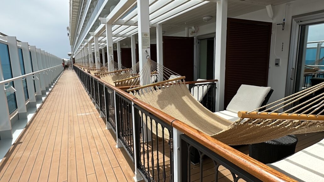 A cruise ship deck on the Carnival Venezia showcases a row of private balconies, each with a hammock and lounge chair. The wood-paneled deck boasts railings and glass panels on the left, while doors lead to sumptuous cabins on the right. The cloudy sky enhances the tranquil setting.