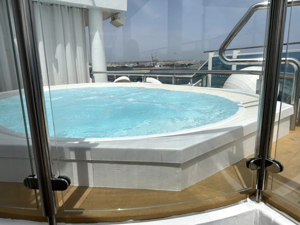 A round jacuzzi sits on a wooden deck surrounded by glass fencing, reminiscent of the serene luxury found in Carnival Venezia cabins. The water is clear and gently rippling, framed by a stunning view of the city and waterfront under a blue sky.